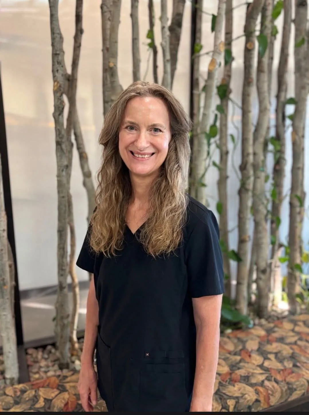 Nancy Orr, smiling, in black scrubs in front of a birch-branch backdrop